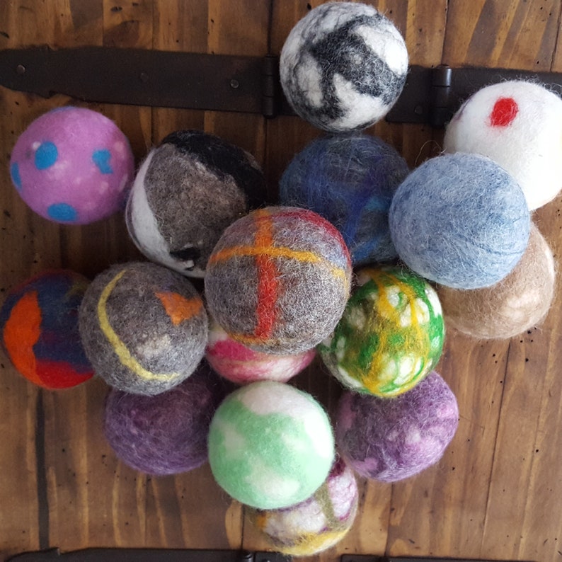 May include: A collection of 14 colorful felt balls, some with patterns, others with solid colors. The balls are arranged in a semi-circle on a wooden surface.