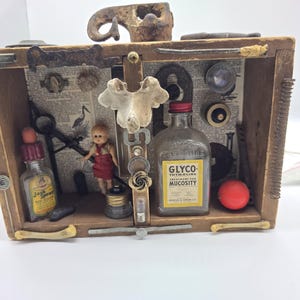 May include: A rectangular wooden shadow box containing a collection of found objects including a small doll in a red dress, a glass bottle labelled Glyco-Thymoline, a Jack Sprat bottle, a bone fragment, metal hardware, and vintage book pages.