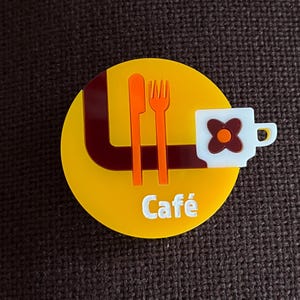 Könnte beinhalten: Gelbes kreisförmiges Café-Logo mit braunem und orangefarbenem Design. Das Logo zeigt eine stilisierte Gabel und ein Messer, eine Kaffeetasse mit Blumenmuster und das Wort "Café" in Weiß.