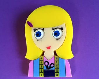 May include: A brooch shaped like a stylized person with bright yellow hair, a purple hair clip, and large blue eyes. The figure wears a pink and blue top featuring a yellow measuring tape graphic with numbers 1 through 14 and a black scissor icon.