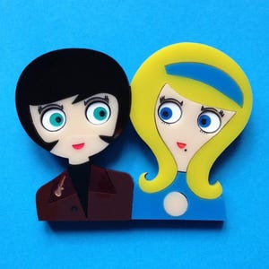 May include: An acrylic brooch featuring two stylized figures. The left figure has black hair, green eyes, and a dark brown jacket. The right figure has yellow hair with a blue headband, blue eyes, and a blue top. Both have pale faces.