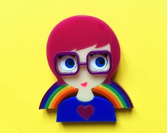 May include: A brooch shaped like a person with bright pink hair, wearing purple-rimmed glasses and a blue shirt with a dark heart graphic. A colorful rainbow arc extends from behind the shoulders, framing the figure against a solid yellow background.