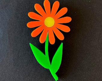 Könnte beinhalten: Eine flache, stilisierte Blumenbrosche mit leuchtend orangefarbenen Blütenblättern, die um eine gelbe Mitte angeordnet sind. Die Blume ist an einem grünen Stiel mit zwei spitzen grünen Blättern befestigt, die von der Basis abstehen.