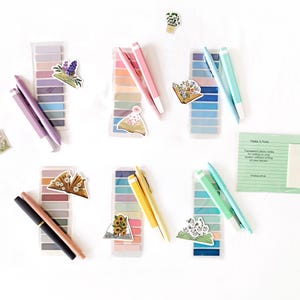 Puede incluir: Seis juegos de pestañas adhesivas transparentes rectangulares en varias paletas de colores, cada uno con dos bolígrafos y una pegatina con temática de libros. Se incluye una tarjeta rectangular verde con el texto Make A Note y notas adhesivas.