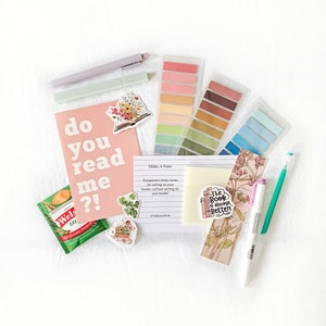 May include: A flat lay of stationery supplies, including a pink notecard with the text "do you read me?!", a pack of colourful sticky notes, a green tea bag, a pen, and a bookmark with the text "The book is always better".