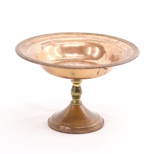 May include: A vintage copper-toned pedestal bowl with a round, shallow dish and a decorative stem. The bowl has a tarnished appearance, with a brass-colored accent on the stem. The base is a wider, rounded shape.