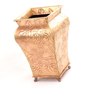 May include: A square brass vase featuring an embossed pattern of grape clusters and vine leaves on all four sides. The vessel has a flared top rim, a tapered body, and stands on four small, decorative metal feet at the base corners.