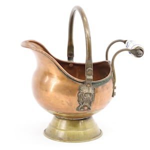 May include: A copper and brass antique sugar scuttle with a handle and spout. The scuttle has a lion's head detail on the front and a ceramic handle with blue and white floral accents. The base is brass.