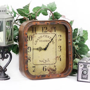 May include: A square, vintage-style clock with a rusty brown metal frame and a handle. The clock face is beige with black numerals and the words "Grand Central Station New York 1913." A decorative lantern and a small camera are also visible.