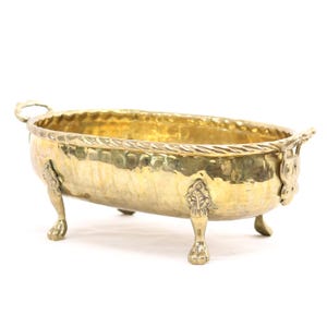May include: A hammered brass oval-shaped decorative bowl with ornate handles and claw feet. The rim features a twisted rope design. The bowl has a golden hue and is set against a white background.