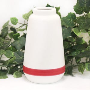 May include: A white ceramic vase with a red band around the base. The vase has a rounded shape and a narrow neck. The background features green foliage.