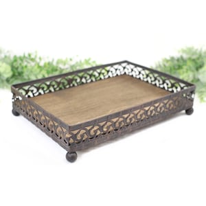 May include: A rectangular tray featuring a dark brown wooden base and a decorative bronze-toned metal rim with an intricate scrollwork pattern. The tray is supported by four small, rounded ball-shaped feet at each corner of the base structure.