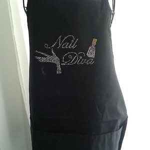 May include: Black apron with the text "Nail Diva" in a cursive font, embellished with rhinestones. The design includes a hand with rhinestones and a nail polish bottle. The apron has two front pockets.