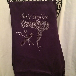 Pu&ograve; includere: Grembiule viola con il testo "hair stylist" in lettere bianche e un design di asciugacapelli e forbici con strass.