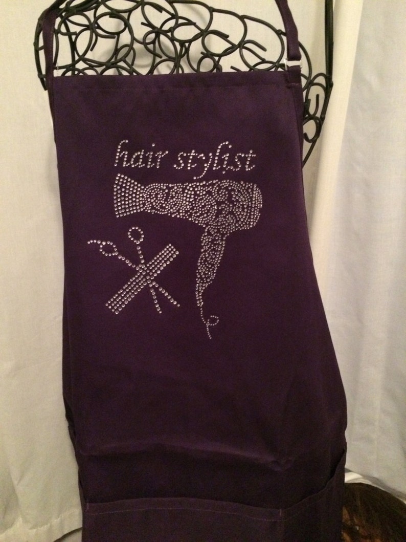 Pu&ograve; includere: Grembiule viola con il testo "hair stylist" in lettere bianche e un design di asciugacapelli e forbici con strass.