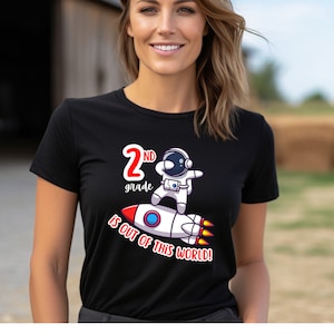 May include: Black t-shirt with a colorful graphic featuring a cartoon astronaut on a rocket ship. The text reads "2nd grade is OUT OF THIS WORLD!" in red and white. The shirt is made of a soft material.