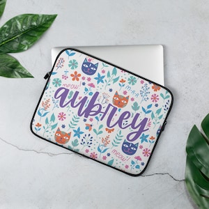 May include: A white laptop sleeve with a colorful floral pattern featuring cats and the name "Aubrey" in purple lettering. The sleeve has a zipper closure and a black trim.