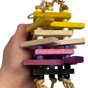 May include: A colourful wooden bird toy with multiple layers of wood and beads. The toy is suspended from a string and has a variety of colours, including yellow, brown, pink, and black.