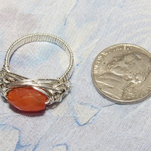 May include: A silver wire wrapped ring with a carnelian gemstone. The ring is shown next to a US quarter for size comparison.