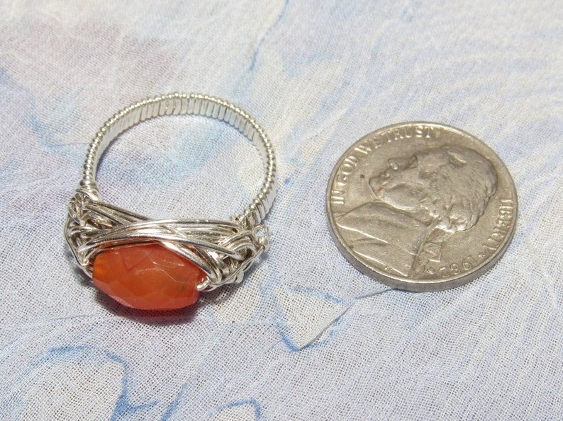 May include: A silver wire wrapped ring with a carnelian gemstone. The ring is shown next to a US quarter for size comparison.