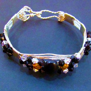 Puede incluir: Un brazalete de alambre plateado con cuentas negras y transparentes, y una cuenta negra grande en el centro.