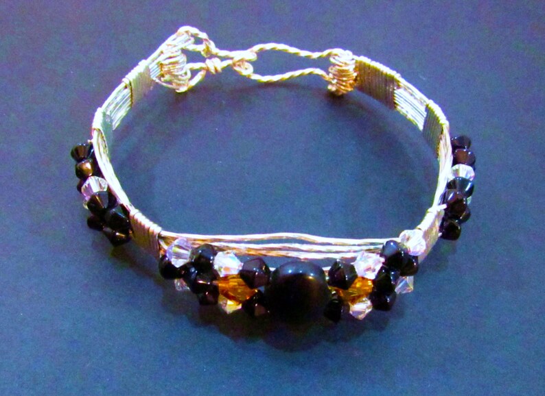 Puede incluir: Un brazalete de alambre plateado con cuentas negras y transparentes, y una cuenta negra grande en el centro.