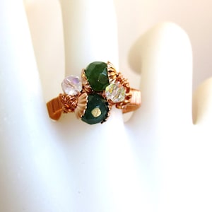 Puede incluir: Un anillo envuelto en alambre de cobre con piedras verdes y transparentes. El anillo est&aacute; sobre un fondo blanco.
