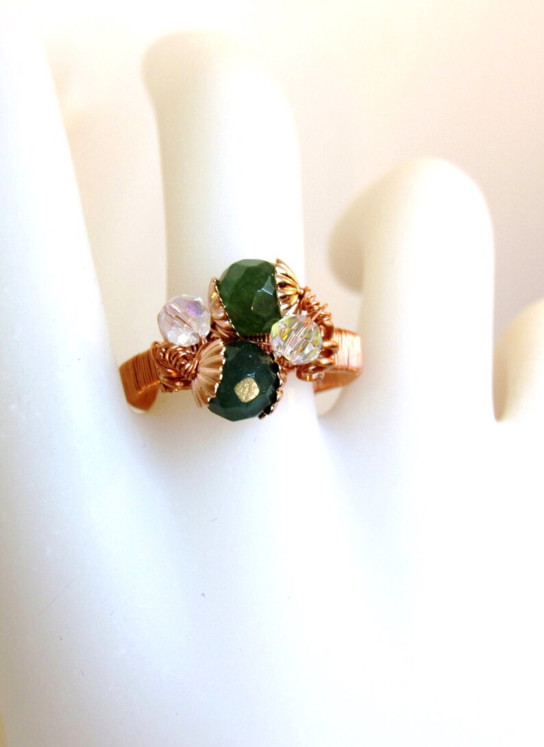 Puede incluir: Un anillo envuelto en alambre de cobre con piedras verdes y transparentes. El anillo est&aacute; sobre un fondo blanco.