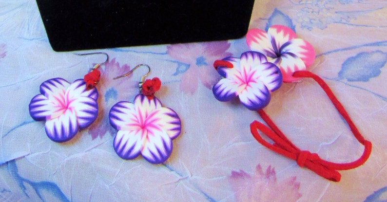 Peut inclure: Un ensemble de boucles d'oreilles et d'un collier assorti en forme de fleurs violettes et blanches. Les fleurs sont en p&acirc;te polym&egrave;re et ont un centre rose. Le collier est fait d'un cordon rouge.