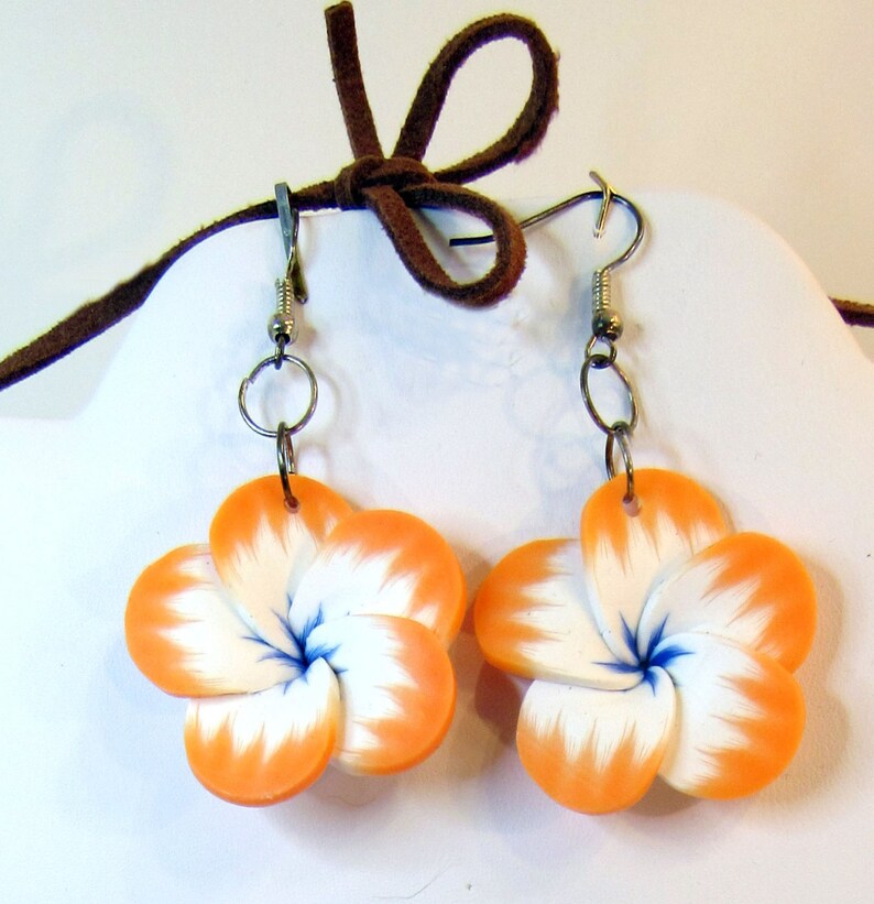 Peut inclure: Une paire de boucles d'oreilles en forme de fleurs orange et blanches avec des centres bleus. Les boucles d'oreilles sont en p&acirc;te polym&egrave;re et ont des crochets argent&eacute;s.