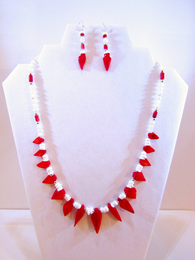 K&ouml;nnte beinhalten: Eine rote und wei&szlig;e Perlenkette mit spitzem Design und passenden Ohrringen. Die Kette und die Ohrringe bestehen aus roten und wei&szlig;en Perlen.