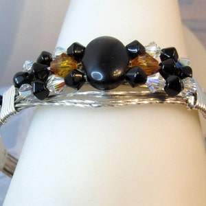 Puede incluir: Un brazalete de alambre plateado envuelto con cuentas negras y &aacute;mbar, y una gran piedra preciosa negra en el centro.