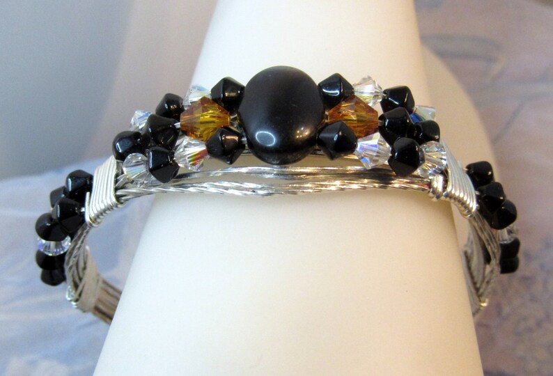 Puede incluir: Un brazalete de alambre plateado envuelto con cuentas negras y &aacute;mbar, y una gran piedra preciosa negra en el centro.