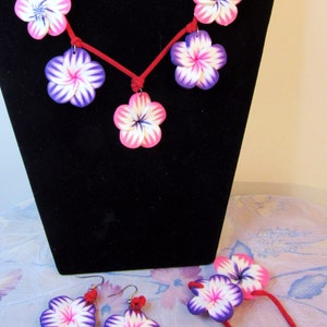 Peut inclure: Un collier en cordon rouge avec cinq breloques en forme de fleurs roses et violettes. Des boucles d'oreilles assorties et un bracelet avec deux breloques en forme de fleurs sont &eacute;galement inclus.