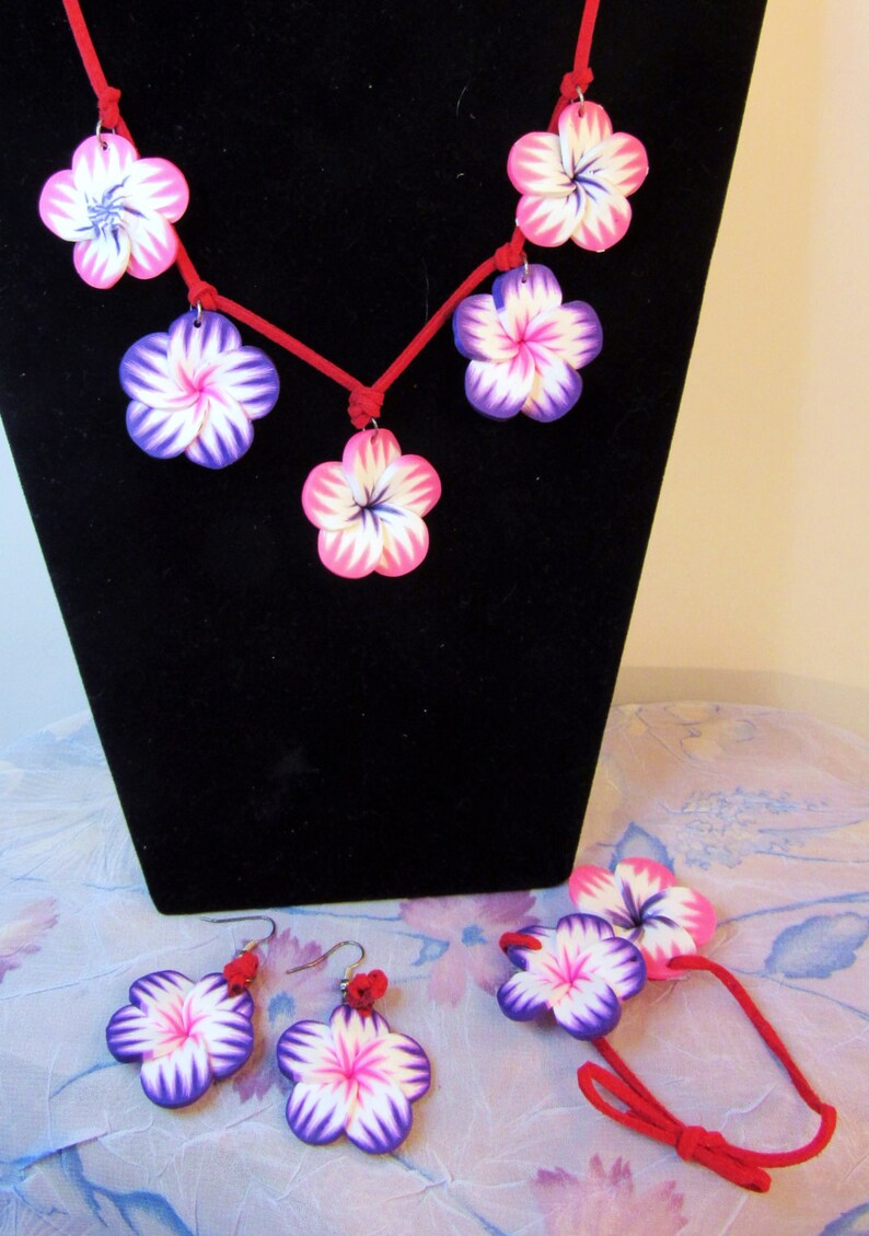 Peut inclure: Un collier en cordon rouge avec cinq breloques en forme de fleurs roses et violettes. Des boucles d'oreilles assorties et un bracelet avec deux breloques en forme de fleurs sont &eacute;galement inclus.