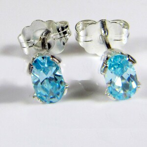Puede incluir: Un par de pendientes de botón con piedras preciosas ovales de color azul claro engastadas en metal plateado. Los pendientes tienen un diseño clásico con cierres de mariposa. Las piedras preciosas son de un color vibrante y llamativo.