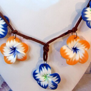 Peut inclure: Un collier en cordon de cuir marron avec cinq breloques en forme de fleurs en p&acirc;te polym&egrave;re. Les fleurs sont blanches avec des accents bleus et orange.