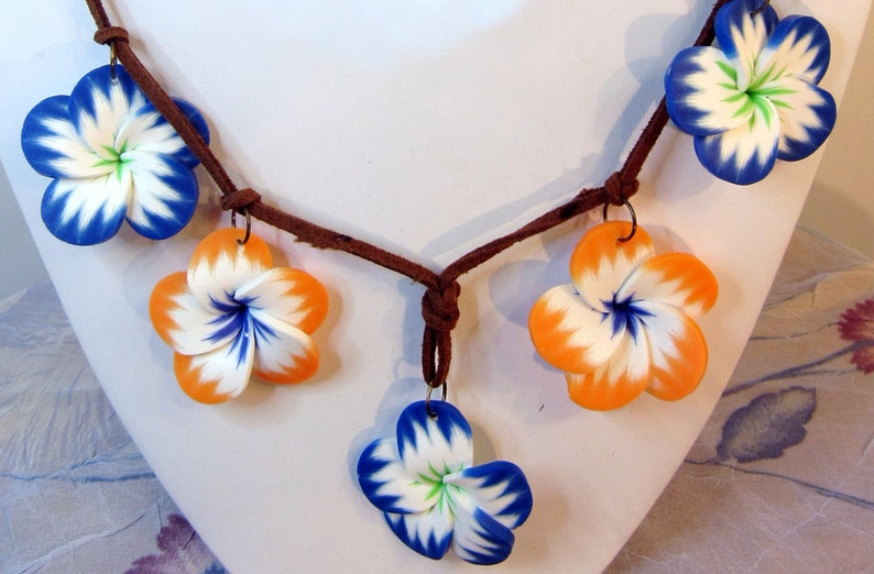 Peut inclure: Un collier en cordon de cuir marron avec cinq breloques en forme de fleurs en p&acirc;te polym&egrave;re. Les fleurs sont blanches avec des accents bleus et orange.