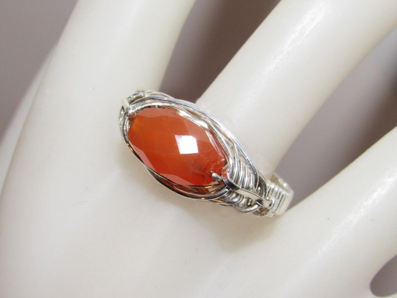 May include: A silver wire wrapped ring with a large, faceted carnelian gemstone. The carnelian is a deep orange color.