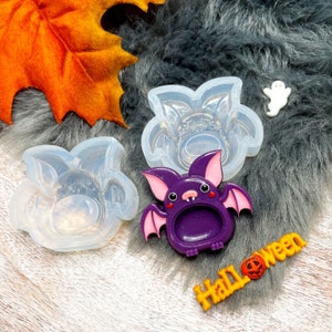 May include: Two clear silicone molds shaped like bats, one with a detailed bat design and the other with a simple bat shape. A purple resin bat with a hollow center is also shown. The word "Halloween" is written in orange letters with a pumpkin design.