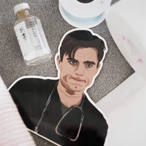 Op de afbeelding: Een gestanste vinylsticker met een gestileerde illustratie van een persoon met donker haar, gekleed in een zwart medisch shirt met een stethoscoop om de nek. De persoon heeft een serieuze gezichtsuitdrukking met een lichte frons.