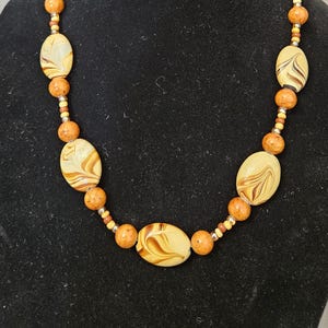 May include: A necklace featuring large, oval-shaped beads with a cream and brown marbled design. The necklace also includes smaller orange and gold-colored beads, strung together to create a unique jewelry piece.