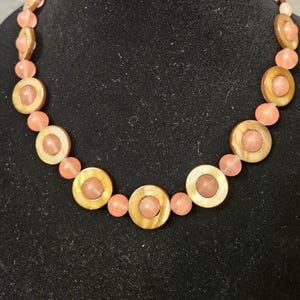 May include: A necklace with alternating round, light brown shell beads and peach-colored round beads. The necklace is displayed on a black mannequin bust.