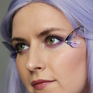 May include: A close-up portrait of a woman with green eyes and lavender hair, featuring butterfly wing eye accents. Her makeup includes purple eyeshadow and a mauve lip colour, creating a fantasy-inspired aesthetic.