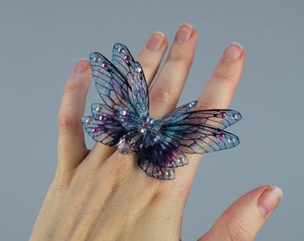 May include: A ring featuring a large, iridescent butterfly with pink and blue wings. The butterfly is made of resin and has a glittery finish. The ring is worn on a person's finger. The text "Just As Strange As I Am" is written above the ring.