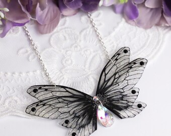 Puede incluir: Un collar con colgante de mariposa con una cadena plateada. La mariposa tiene alas negras y blancas con un centro brillante e iridiscente. El collar está sobre un fondo de encaje blanco con flores moradas.