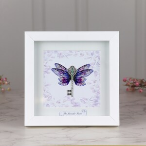 May include: A white framed print of a purple and blue butterfly with a silver key in the centre. The background is a light purple with a floral pattern. The text "The Lavender Fairy" is at the bottom of the print.