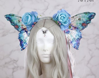 May include: A white headband with two blue butterfly wings and two blue roses on each side. The headband is decorated with silver and crystal accents. The text "Just As Strange As I Am" is visible in the top right corner of the image.