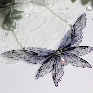May include: A delicate silver chain necklace featuring a large, iridescent, purple butterfly pendant. The butterfly wings are translucent and have a detailed vein pattern. The pendant is accented with small, clear rhinestones.