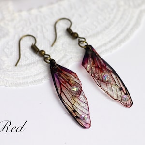 May include: A pair of earrings featuring translucent wings with a red, purple, and black color scheme. The wings have a delicate, intricate design and are accented with small, iridescent rhinestones.